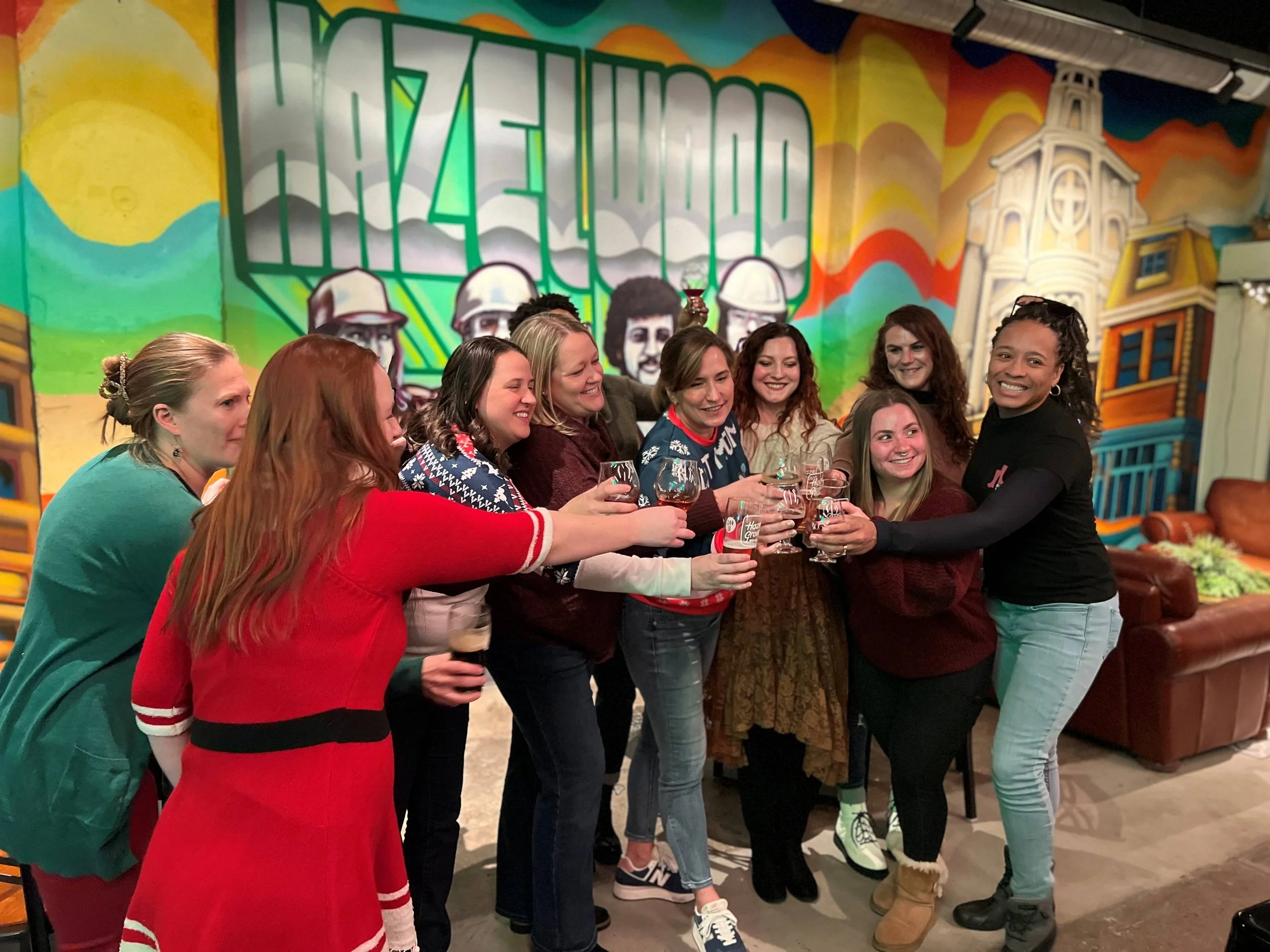 Group of women celebrating with craft beer in front of the vibrant mural art in the Hazel Grove taproom in Pittsburgh.