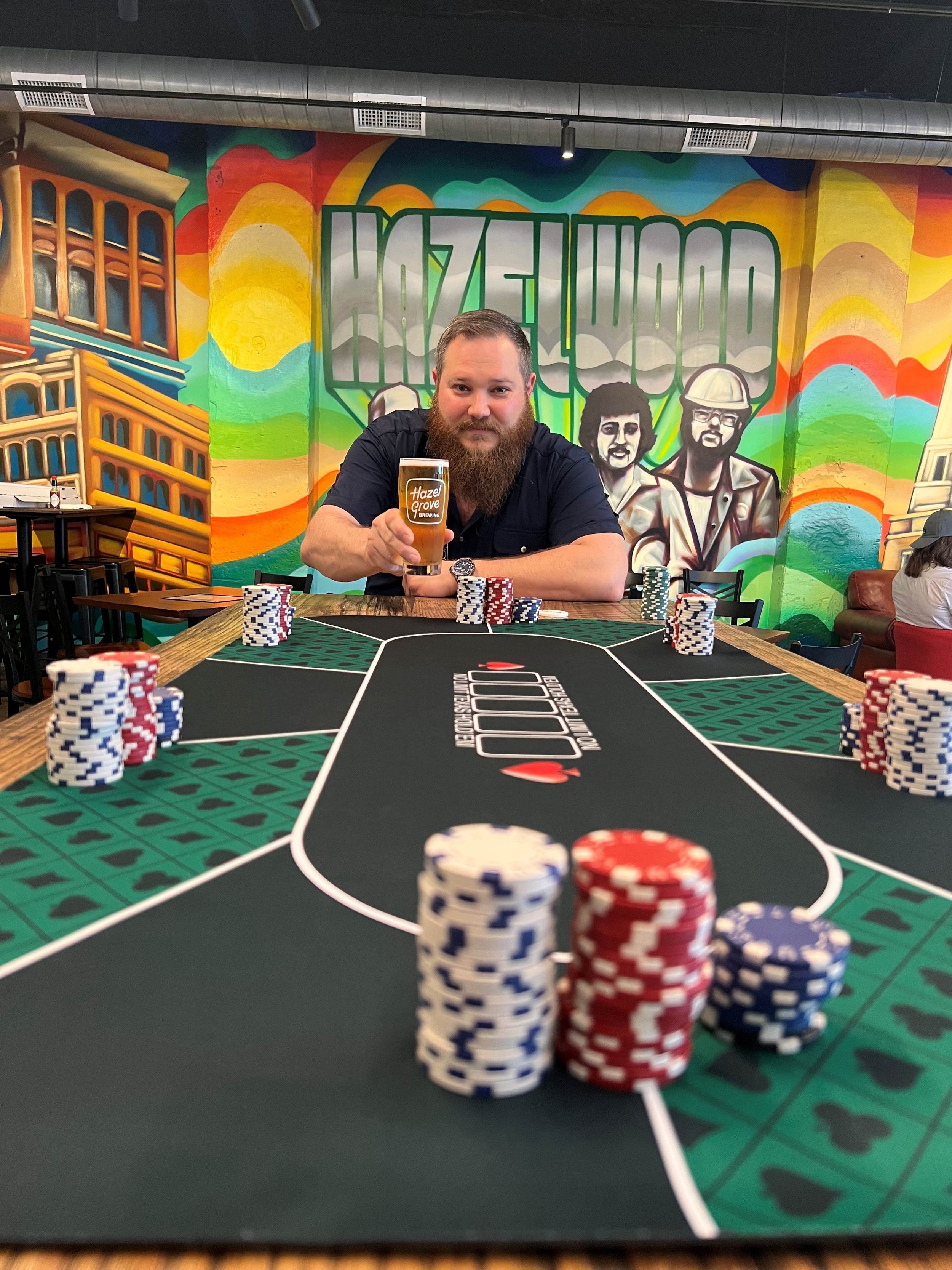 A man with a beard hosting a poker tournament in the Hazel Grove Brewing taproom holding a glass of craft beer with colorful mural art in the background.