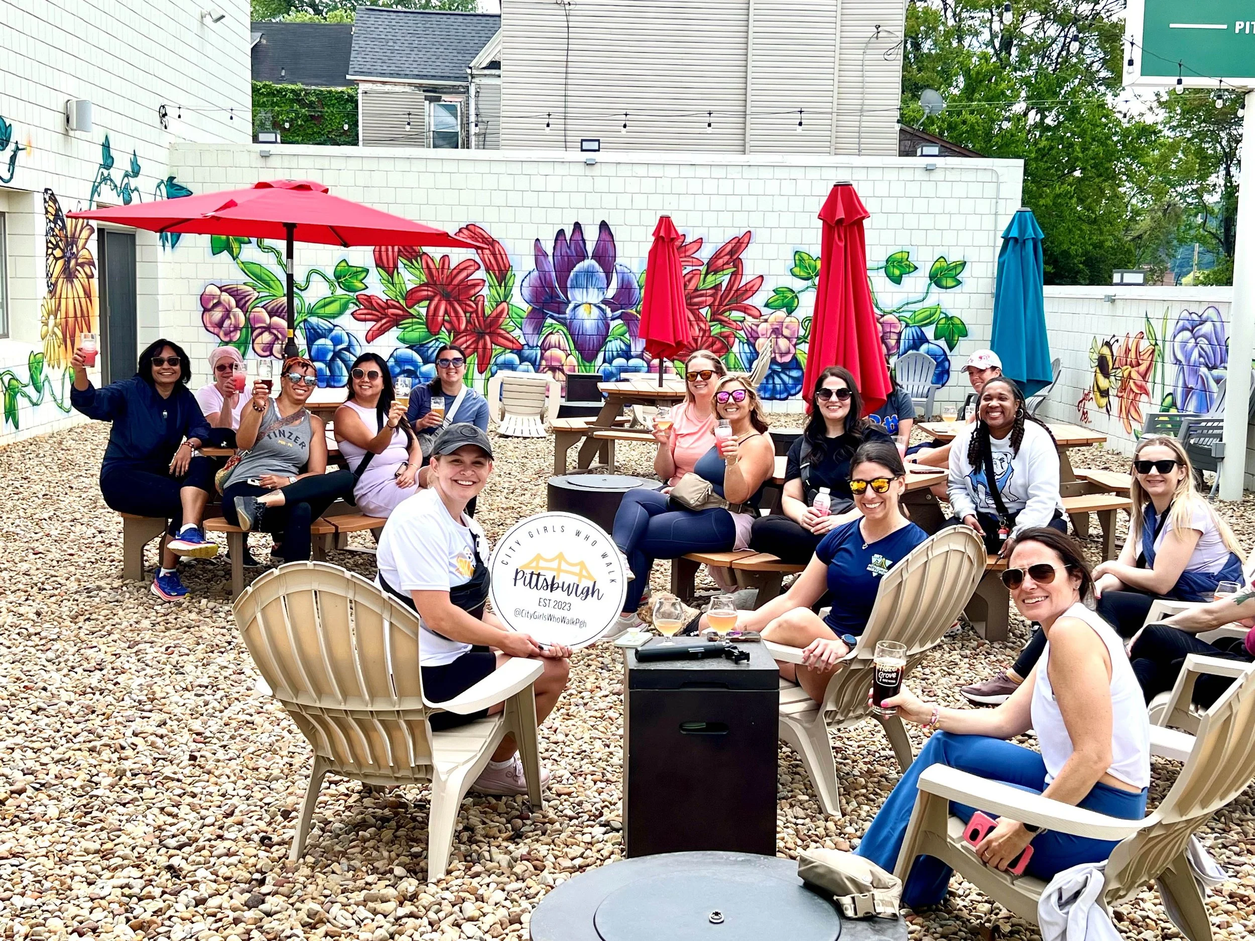 Group of women enjoying craft beer at an outdoor gathering in the Hazel Grove Brewing courtyard with colorful umbrella shades, comfortable seating, and a mural with vibrant flowers on a white brick wall in the background.