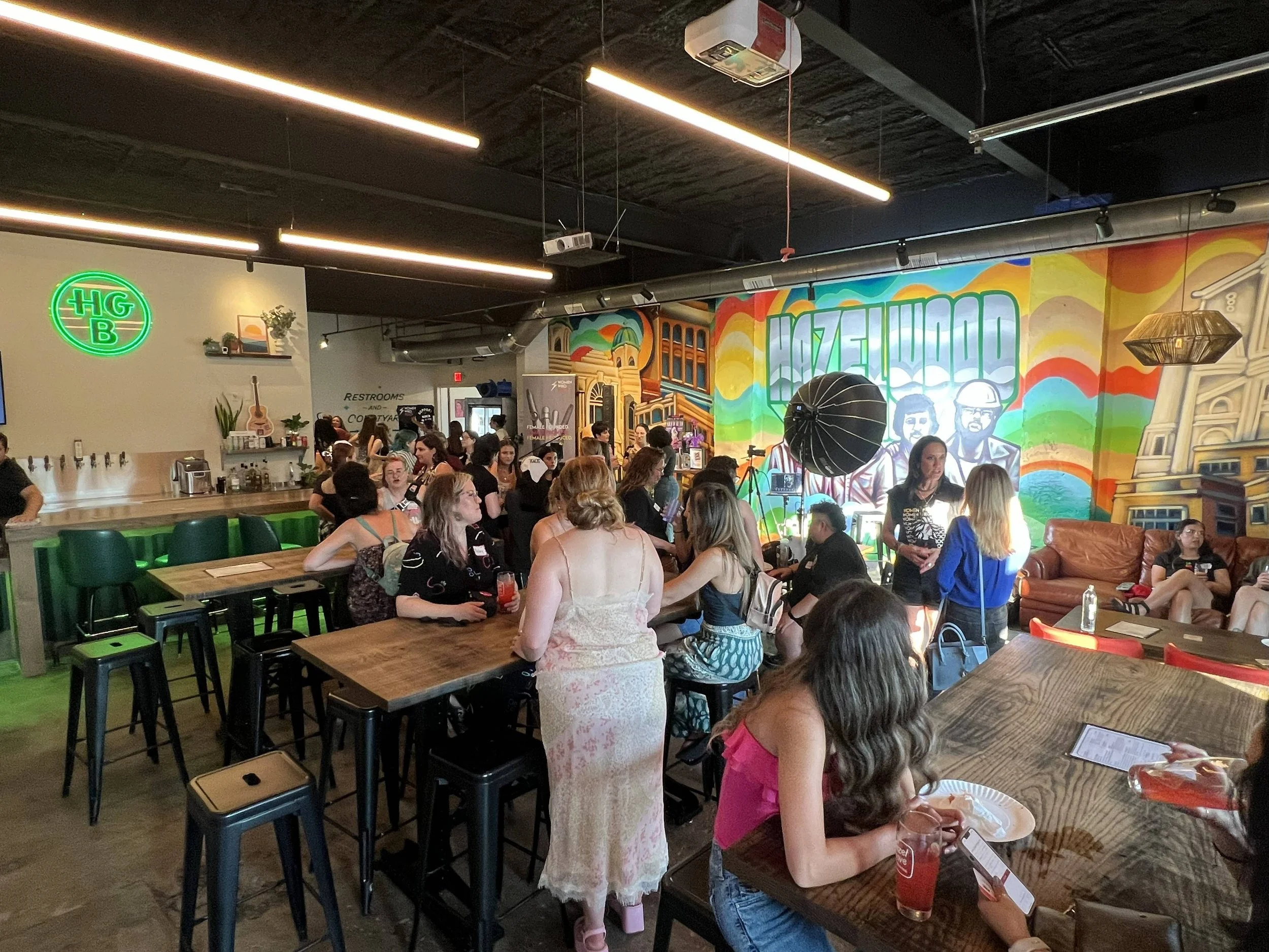 People gathered for a private event in the Hazel Grove Brewing taproom in Pittsburgh in front of a colorful mural on the wall, high tables, and seating. The setting includes neon signs, musical instruments, and party lighting.