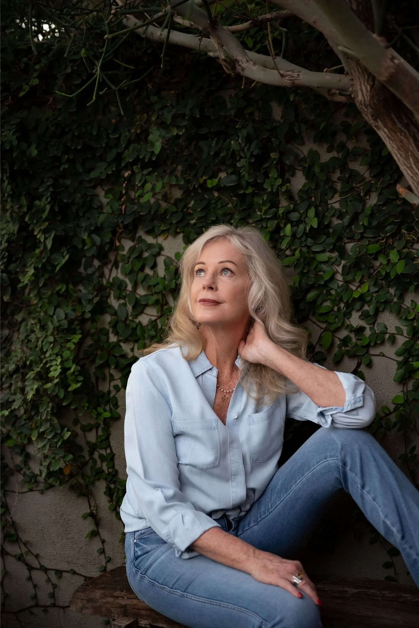 A middle-aged woman with long, wavy blond hair, wearing a light blue denim shirt and jeans, sitting on a wooden bench outdoors against a backdrop of dark green climbing plants and branches, gazing slightly upward.