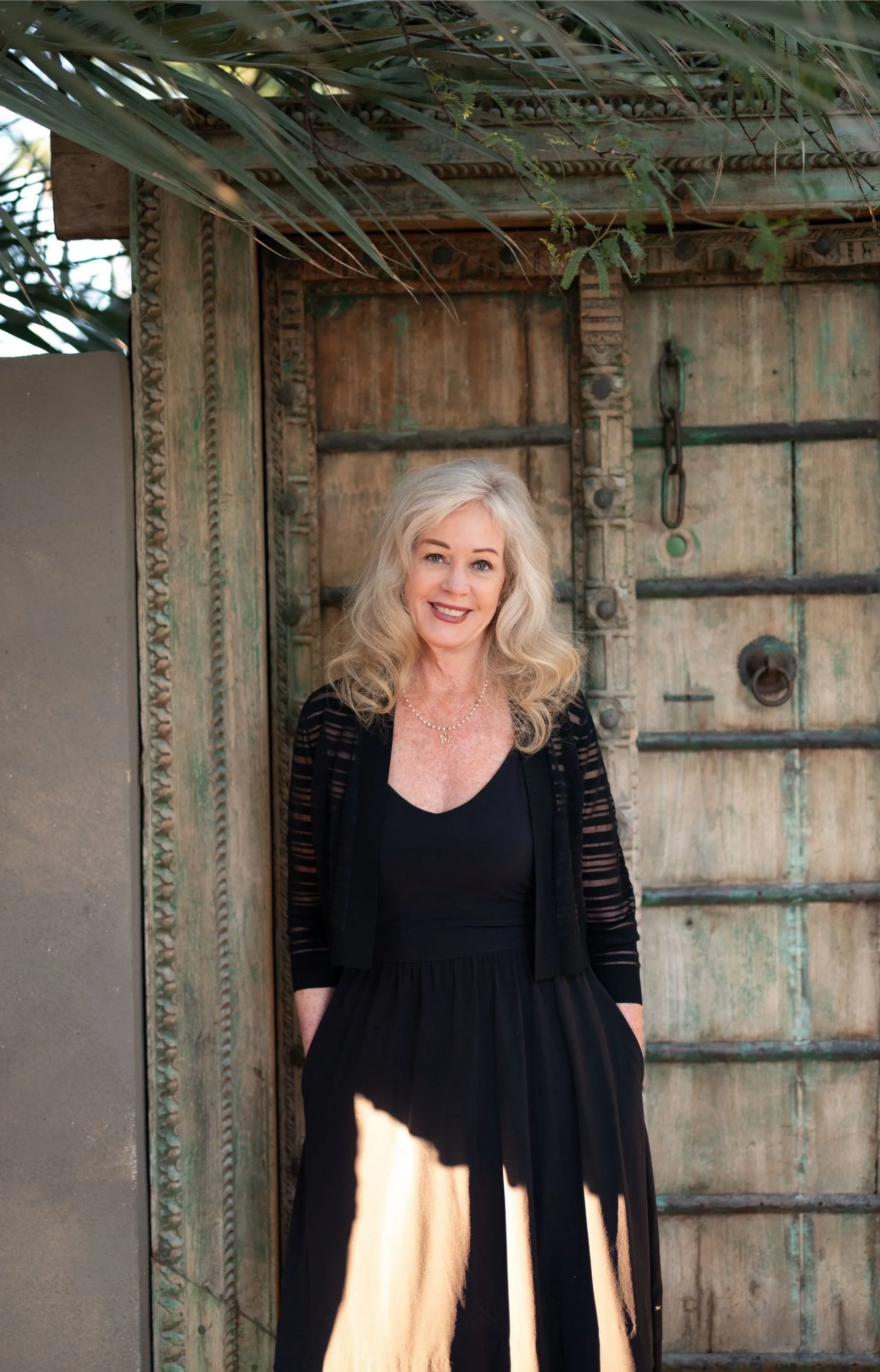 A woman with blonde wavy hair, wearing a black dress and a black sheer cardigan, stands smiling in front of an ornate wooden door with metal accents and greenery overhead.