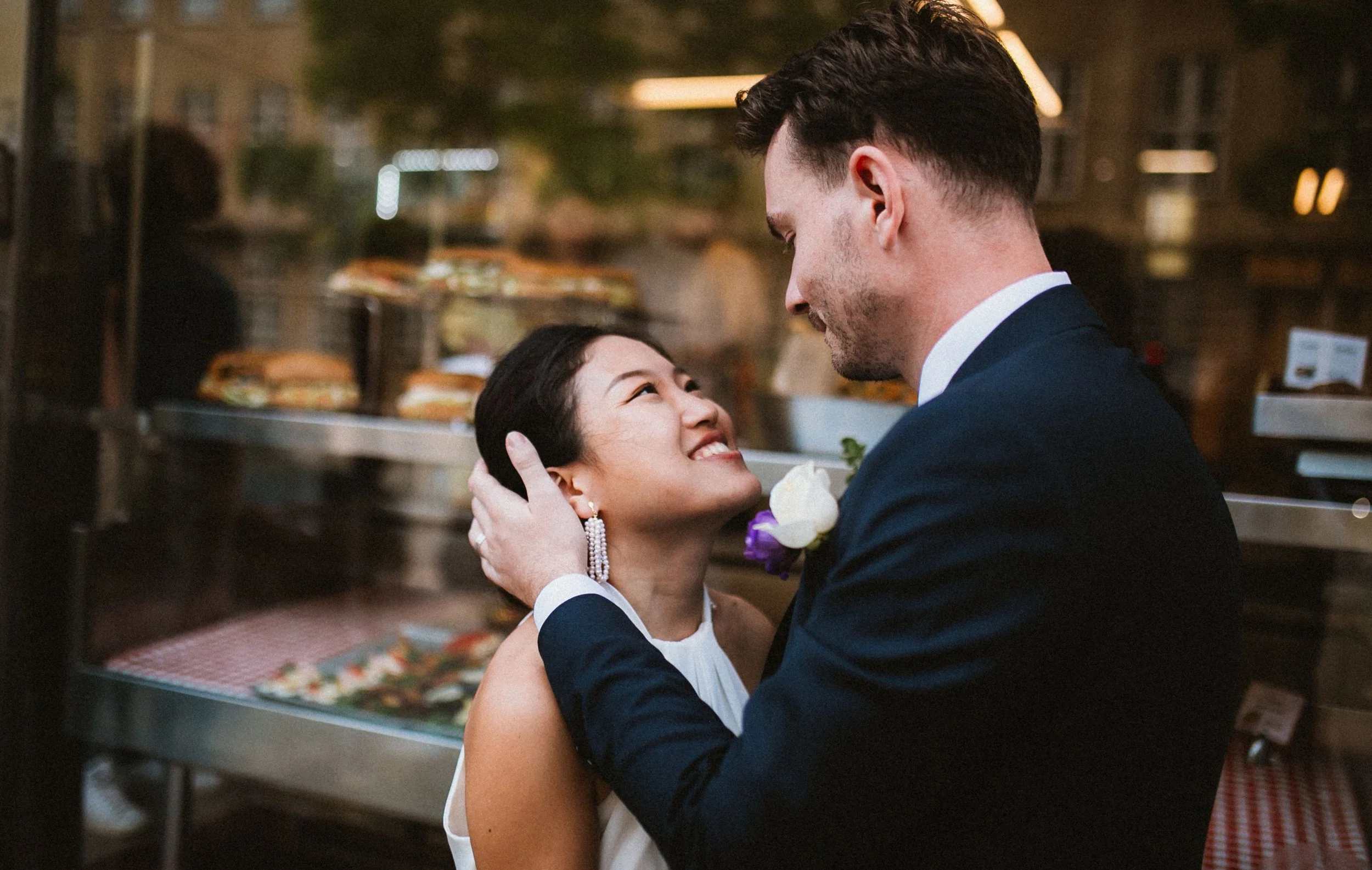 A couple on their wedding day in a bakery, the bride looking up at the groom who is holding her face gently with both hands, smiling.
