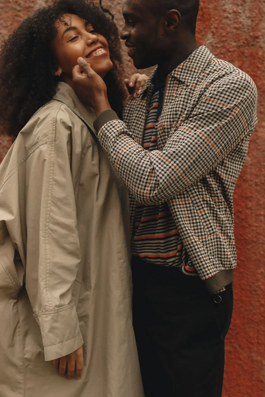 A man and woman are close together, smiling and touching faces. The woman has curly hair and is wearing a beige coat. The man has short hair and is wearing a checkered jacket over a striped shirt. They are standing against a red, textured wall.