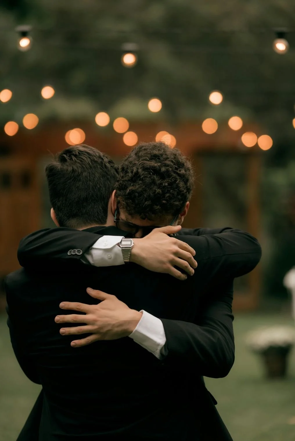 Two men in suits hugging each other warmly, with blurred warm string lights in the background.