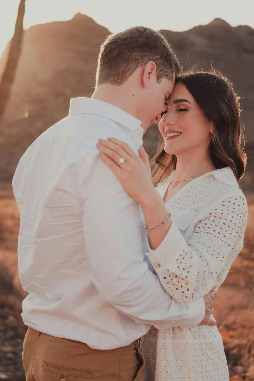 A happy couple embracing outdoors during sunset, with mountains in the background. The woman has dark hair and wears a white eyelet dress and jewelry, while the man has short light brown hair and wears a white shirt and tan pants.