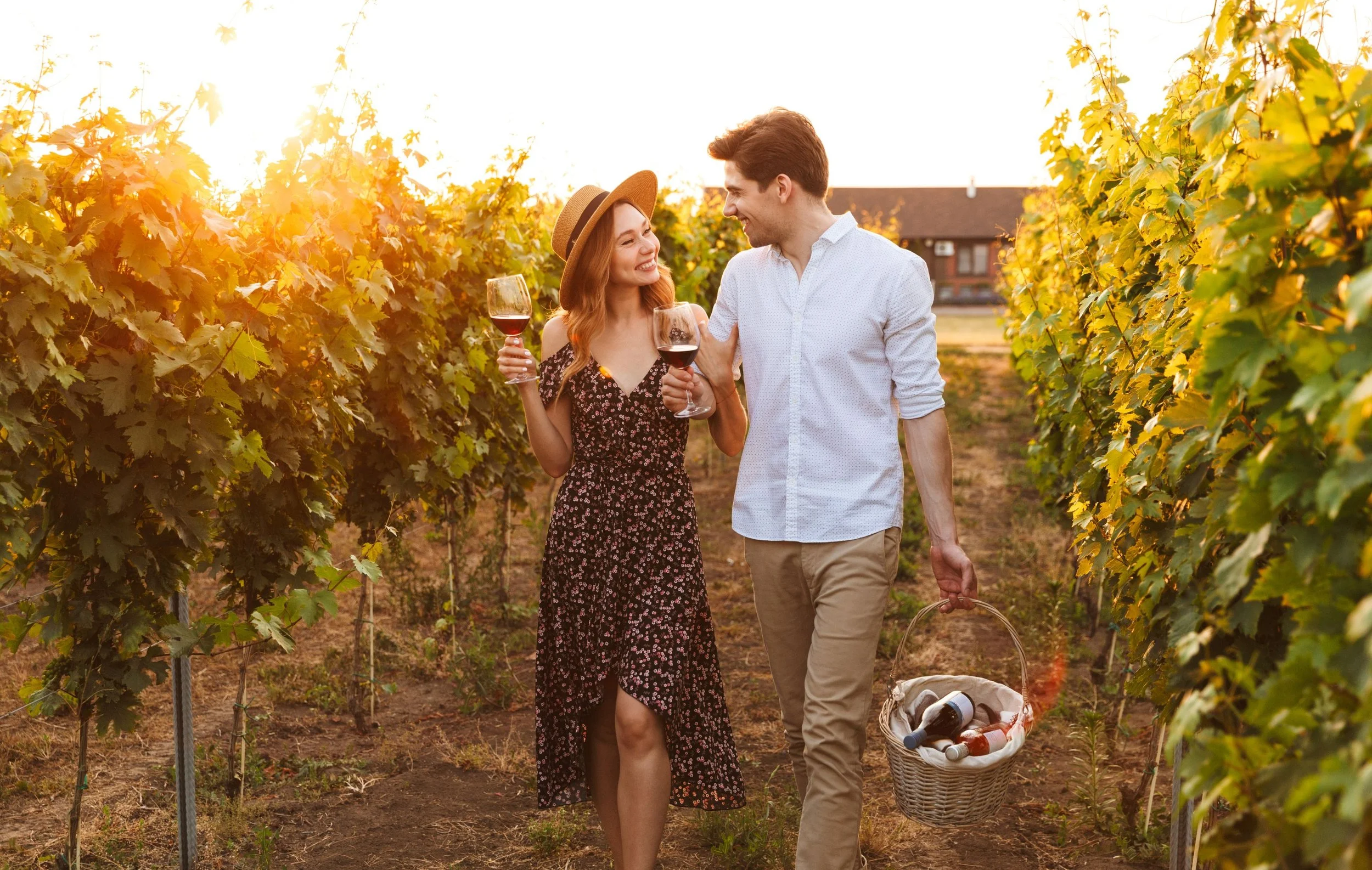 A smiling couple walking through a vineyard during sunset, holding glasses of red wine, with the man carrying a basket of wine bottles.