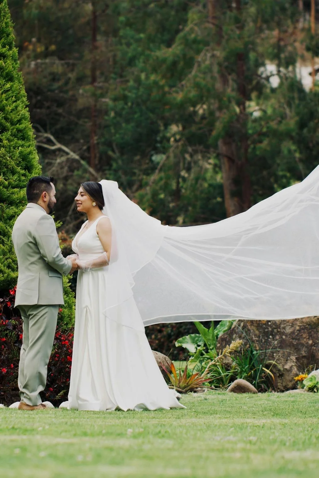A bride and groom holding hands outdoors with trees in the background, the bride in a white gown and veil, and the groom in a light-colored suit, during their wedding ceremony.