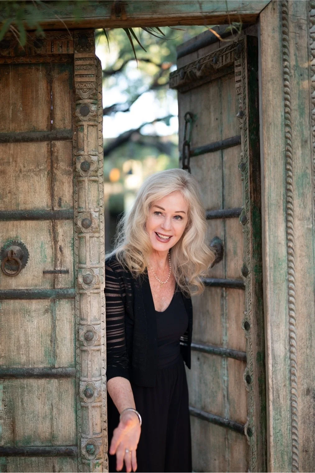 A smiling woman with long curly blonde hair, wearing a black dress and jewelry, peeks out from an ornately carved wooden doorway outdoors.