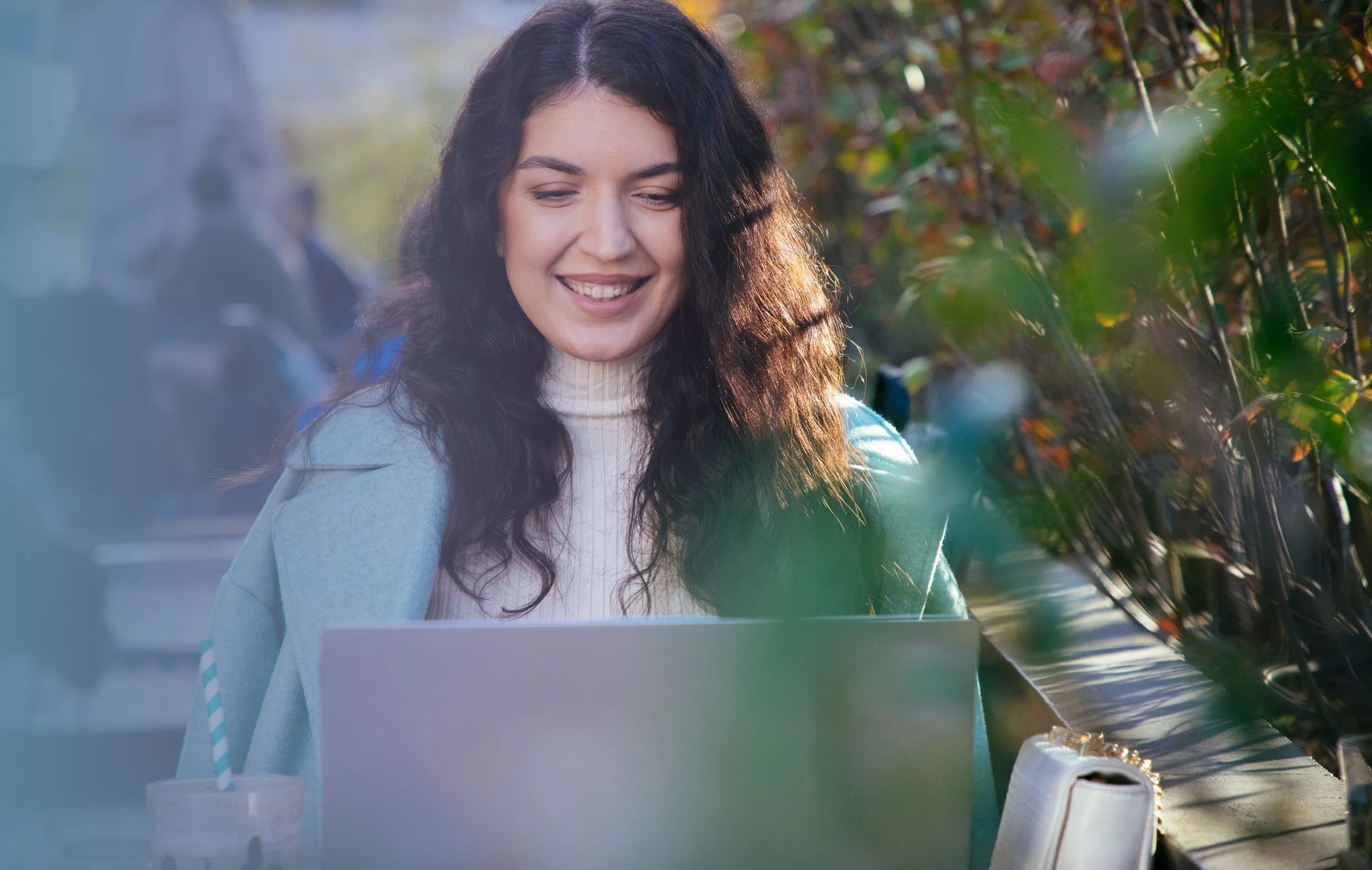 A woman with long curly brown hair smiling while working on a laptop at an outdoor café during daytime.