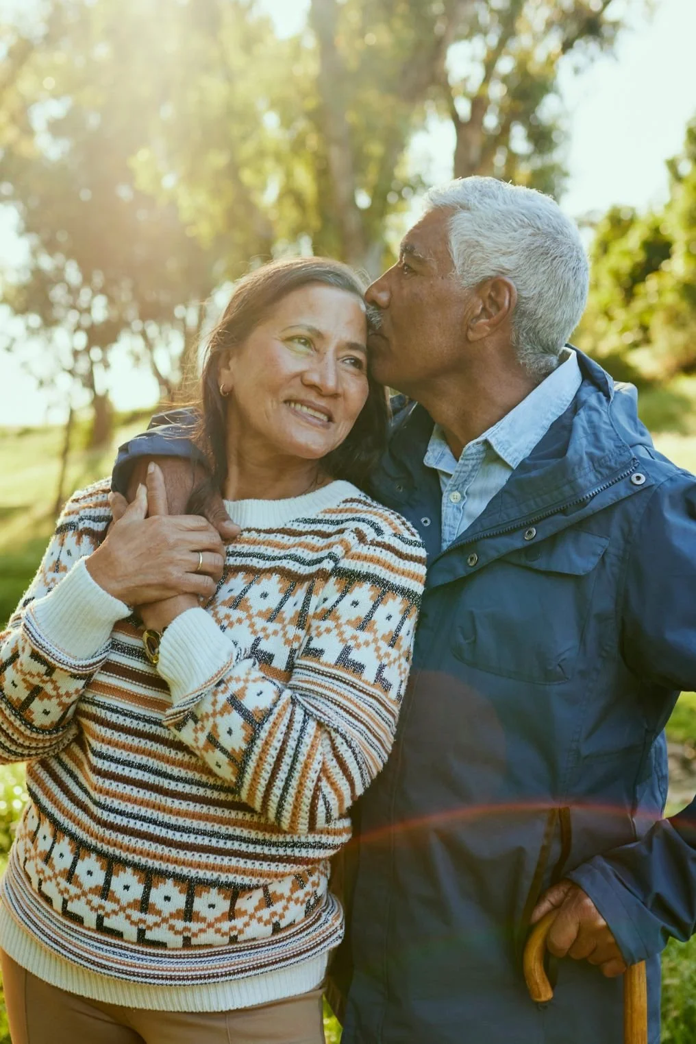 An elderly couple outdoors, with the man kissing the woman on the forehead while she smiles. The man has white hair and holds a cane, and they are surrounded by trees with sunlight filtering through.
