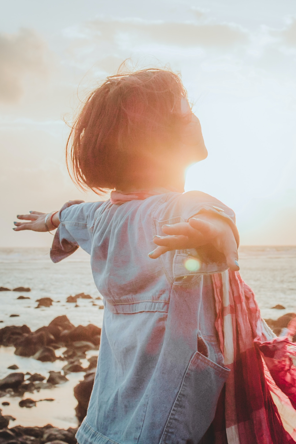 A woman with short hair standing on a rocky beach during sunset, with her arms outstretched and the sun behind her, creating a backlit effect and a lens flare.