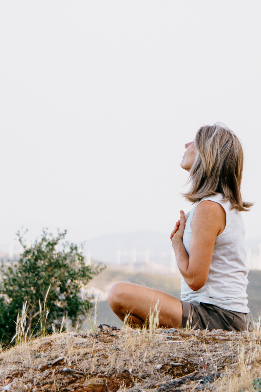 A woman practicing yoga outdoors in a seated meditation pose on a grassy hill with wind turbines in the background.