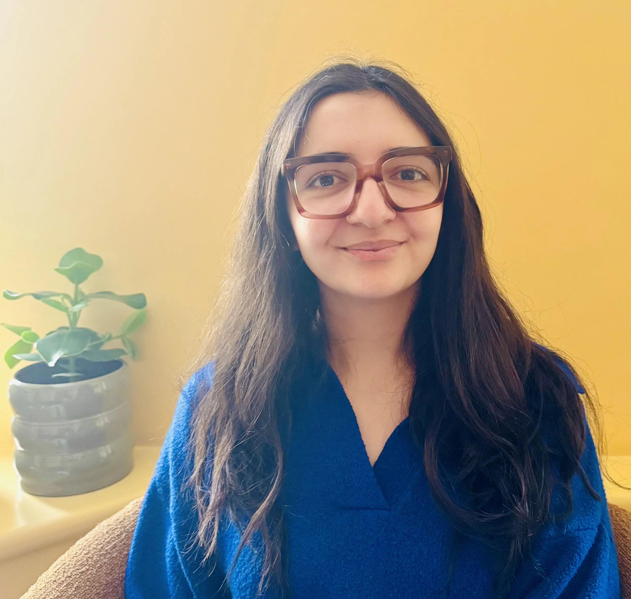 A woman with long dark hair wearing glasses and a blue top, smiling in front of a yellow wall with a plant in a pot on a side table.