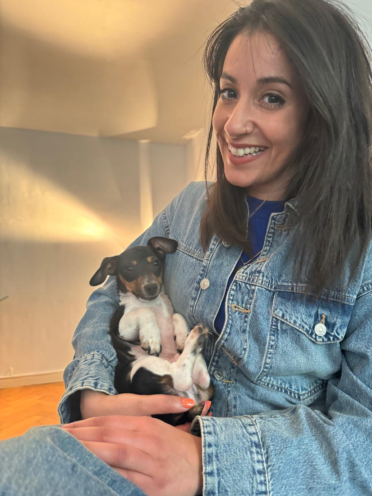 A woman with dark hair and a denim jacket holding a small black and tan puppy, smiling indoors.