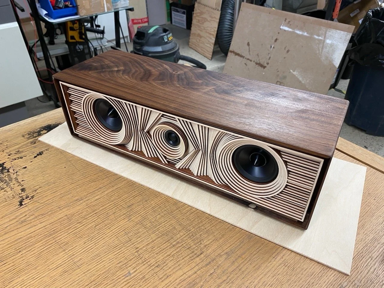 Wooden speaker with circular patterns on the front and three black drivers, placed on a wood table in a workshop.
