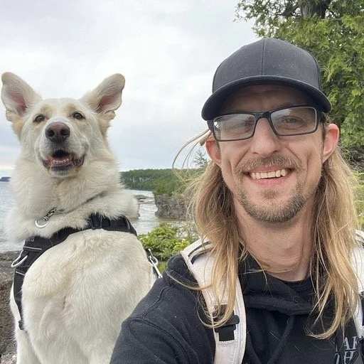 A man with long hair, glasses, wearing a black cap and jacket, taking a selfie with a smiling white dog wearing a harness outdoors near water and trees.