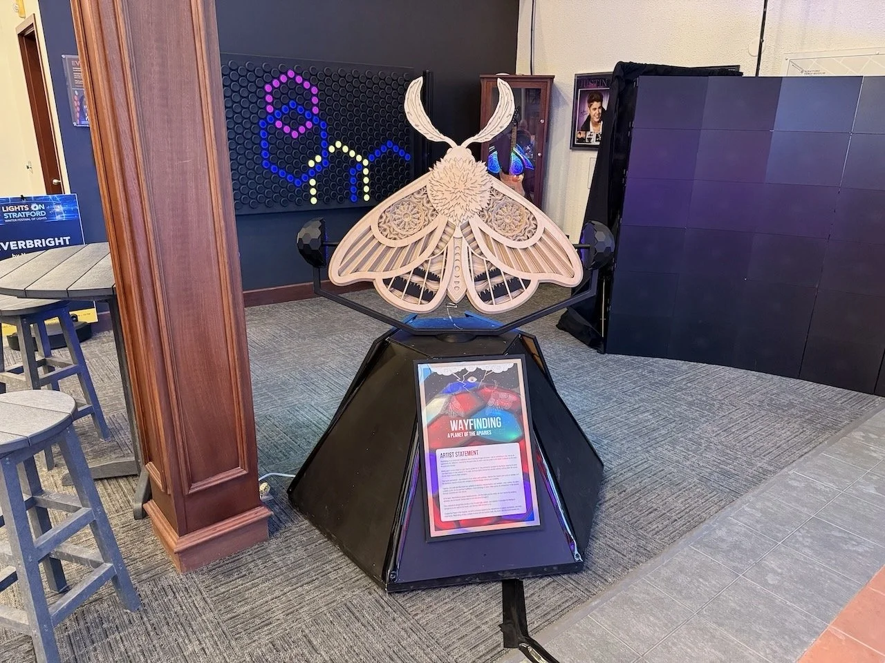 Wooden butterfly sculpture displayed on a black pyramid-shaped stand with an artist statement plaque, located indoors near a wooden pillar and a gray carpeted floor.