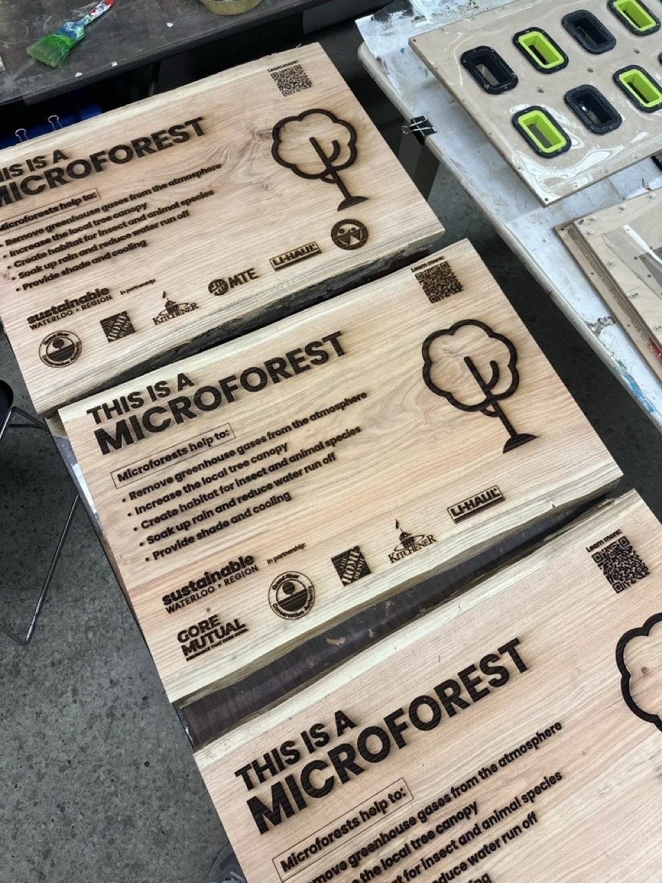 Wooden signs with text about microforests and their benefits, featuring a logo with a tree icon, QR codes, and various sponsor logos, placed on tables.