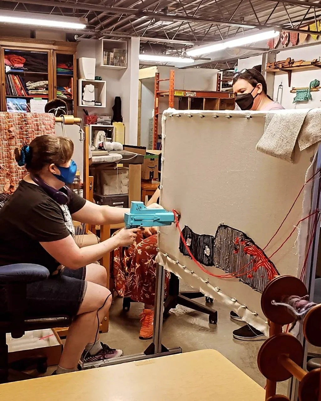 Person wearing a black mask holding a blue toy gun, aiming at a makeshift target on a fabric screen, in a workshop or craft space.