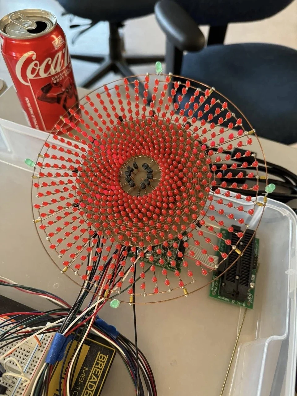 A large circular LED display made of red LEDs on a wire frame, connected to electronic components and wires, with a can of Coca-Cola and a chair in the background.