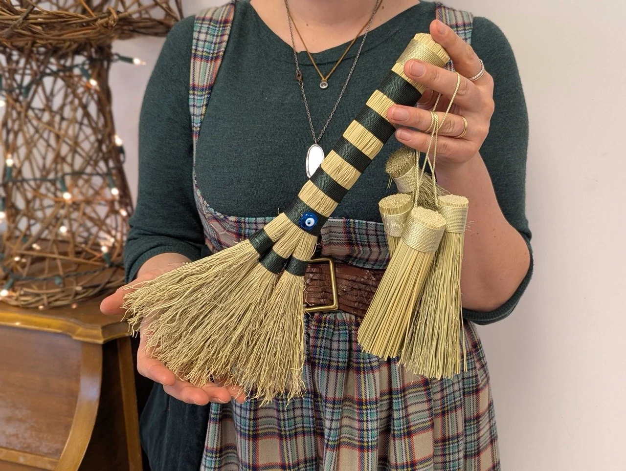 A person holding a decorative broom or duster with gold and black accents, hung with small straw tassels. The person wears layered necklaces, a green top, and plaid pants with a brown belt. There is a wicker basket or structure with lights in the bac