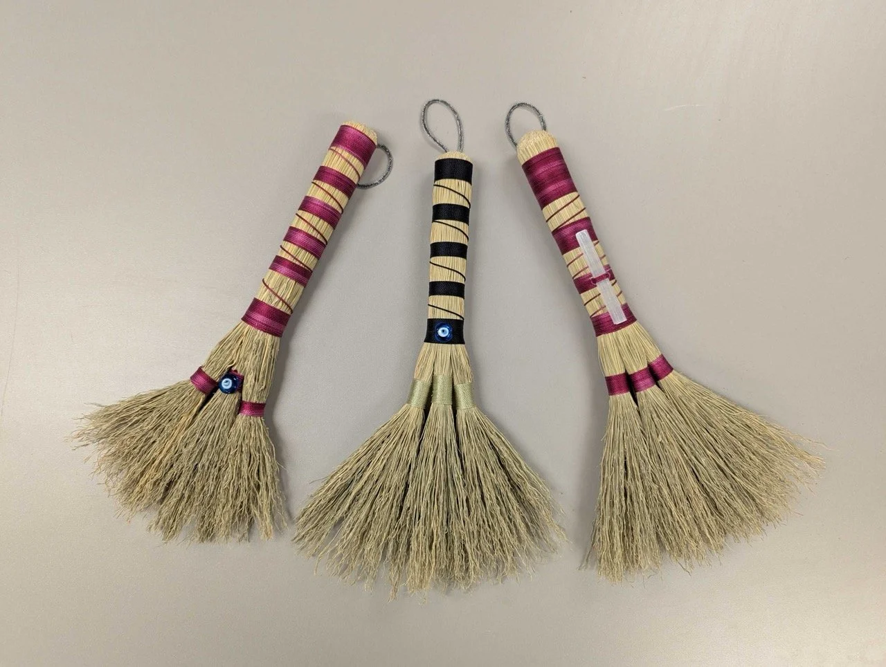 Three handmade broomlets with bamboo handles, wrapped with purple and black ribbon, and decorated with beads, arranged in a row on a light-colored surface.