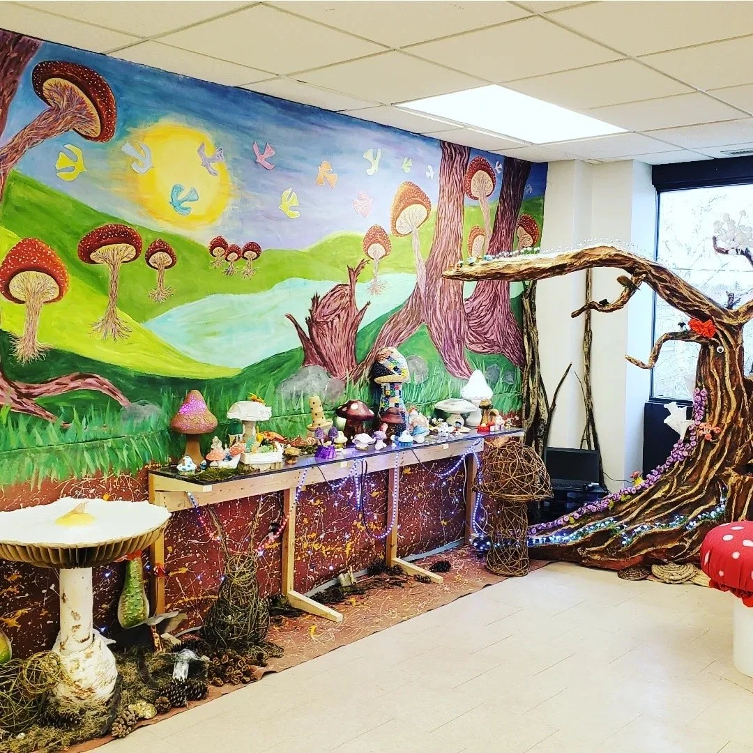 Colorful fantasy-themed room with mushroom and tree decorations, mushroom-shaped lamps, and a painted mural of a whimsical forest scene with a bright sun and flying birds.