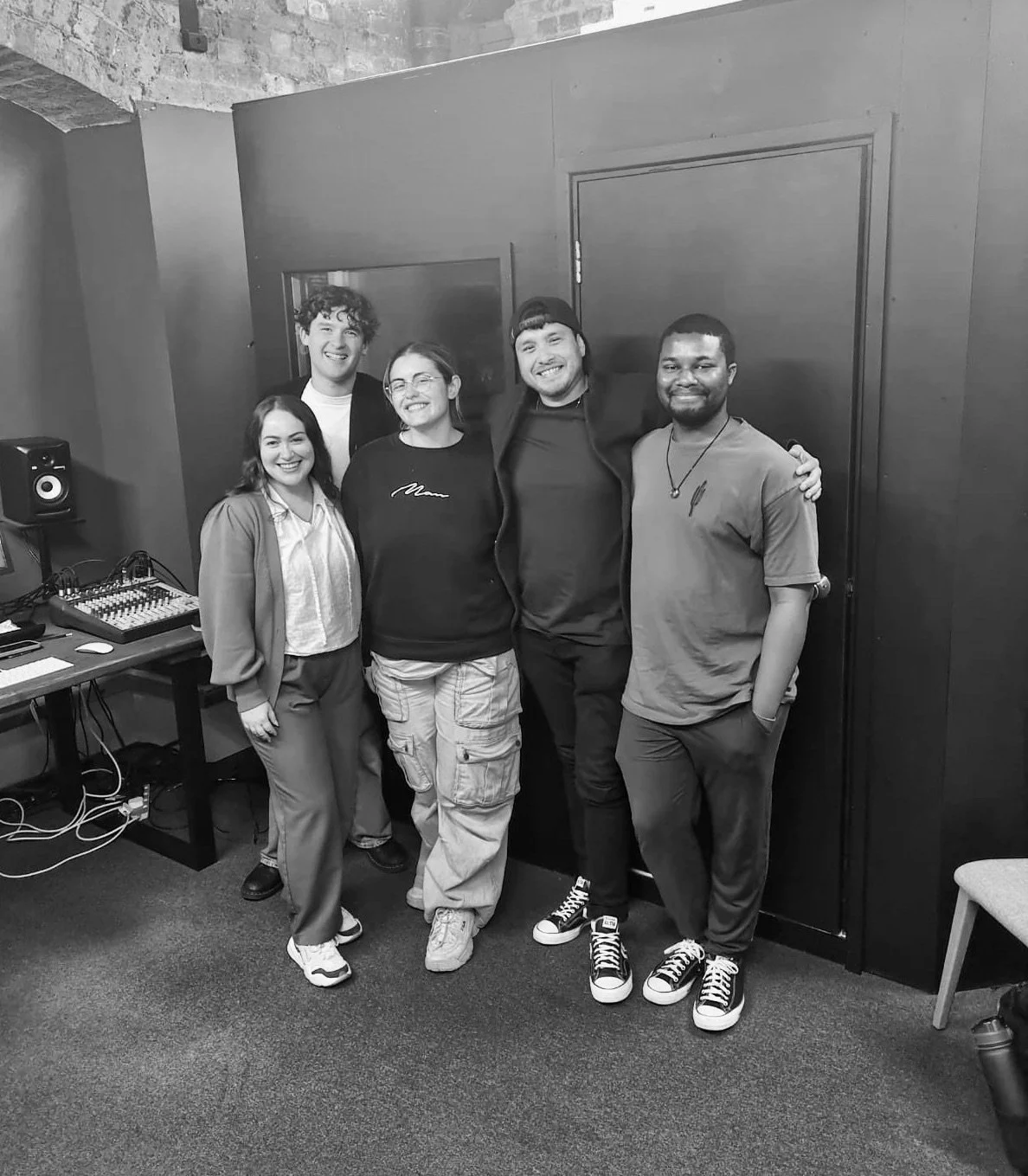 Group of five young voice actors smiling and posing in a recording studio with audio equipment and a computer in the background.