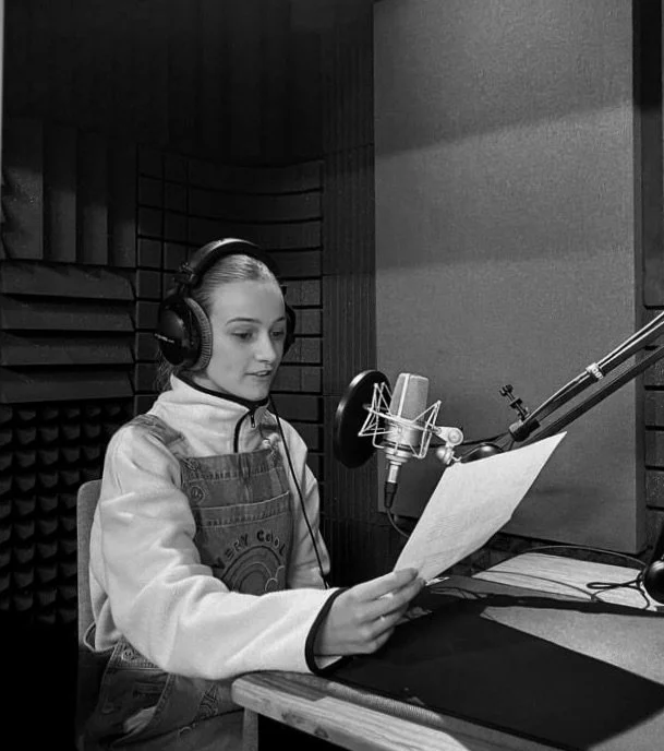 Young woman in a recording studio, wearing headphones, reading a script into a microphone.