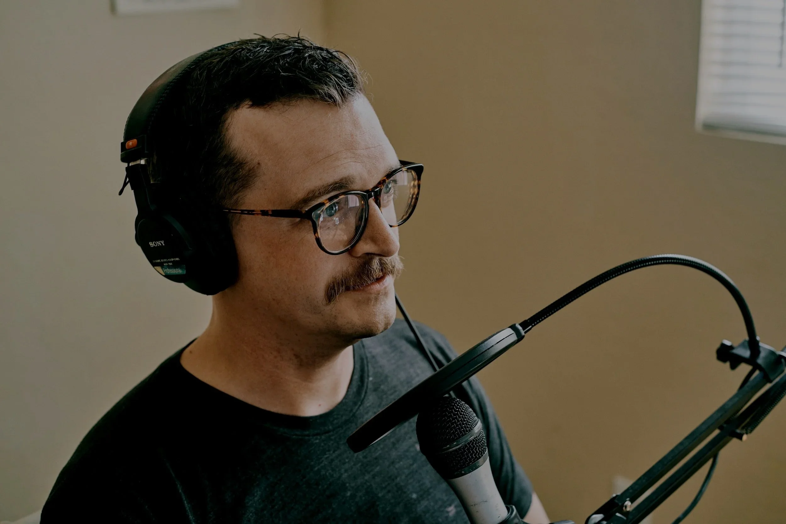 Man with glasses and mustache wearing headphones, speaking into a microphone.