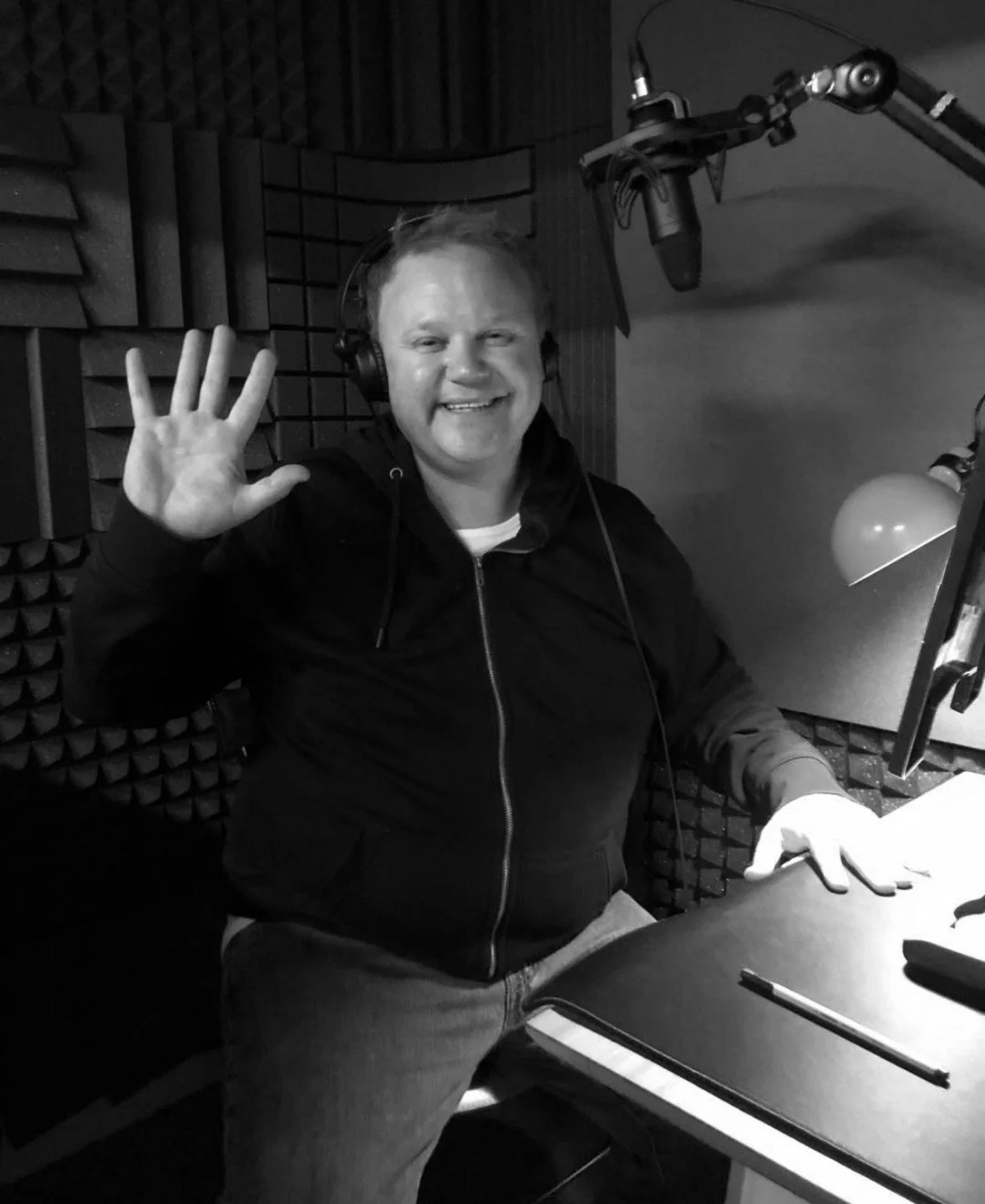 A smiling man in headphones waving in a recording studio with soundproofing panels.