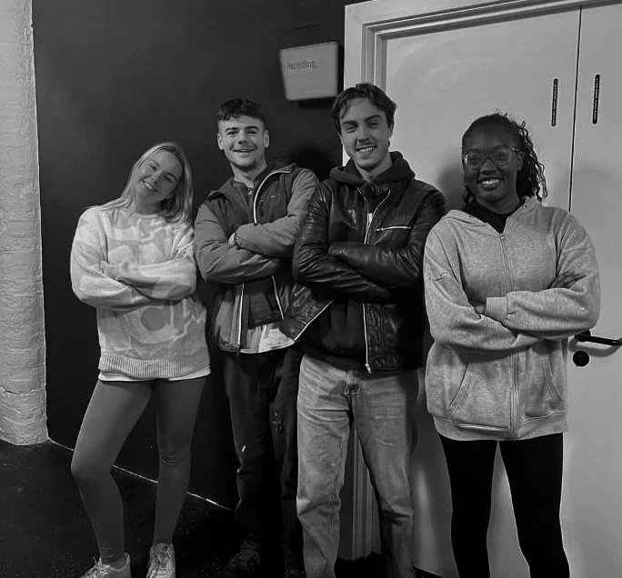 Four young people standing side by side with arms crossed, smiling at the camera in a voiceover studio in Manchester