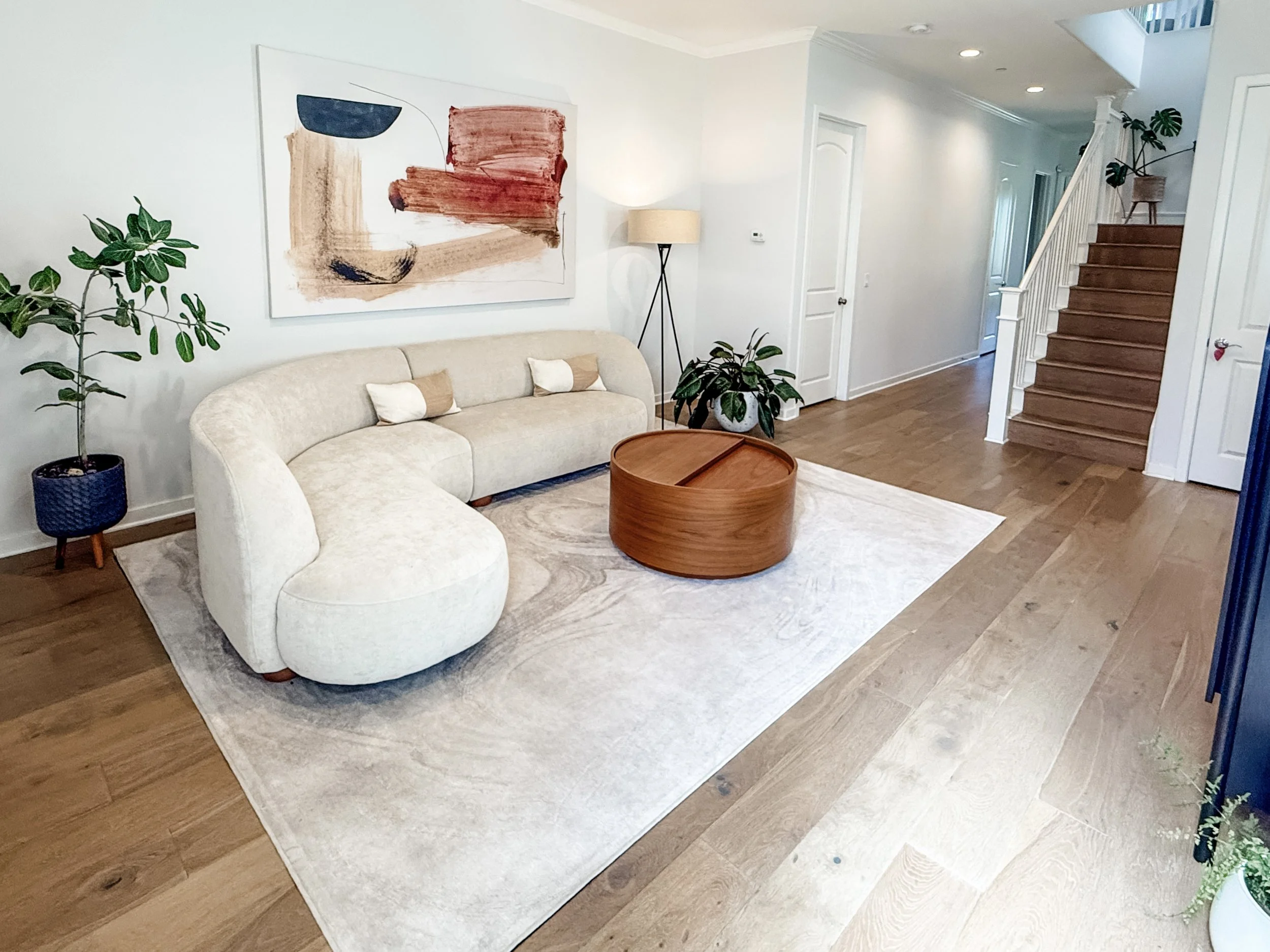 Living room with a beige sofa, green indoor plants, an abstract painting, and a wood stairs leading upstairs.