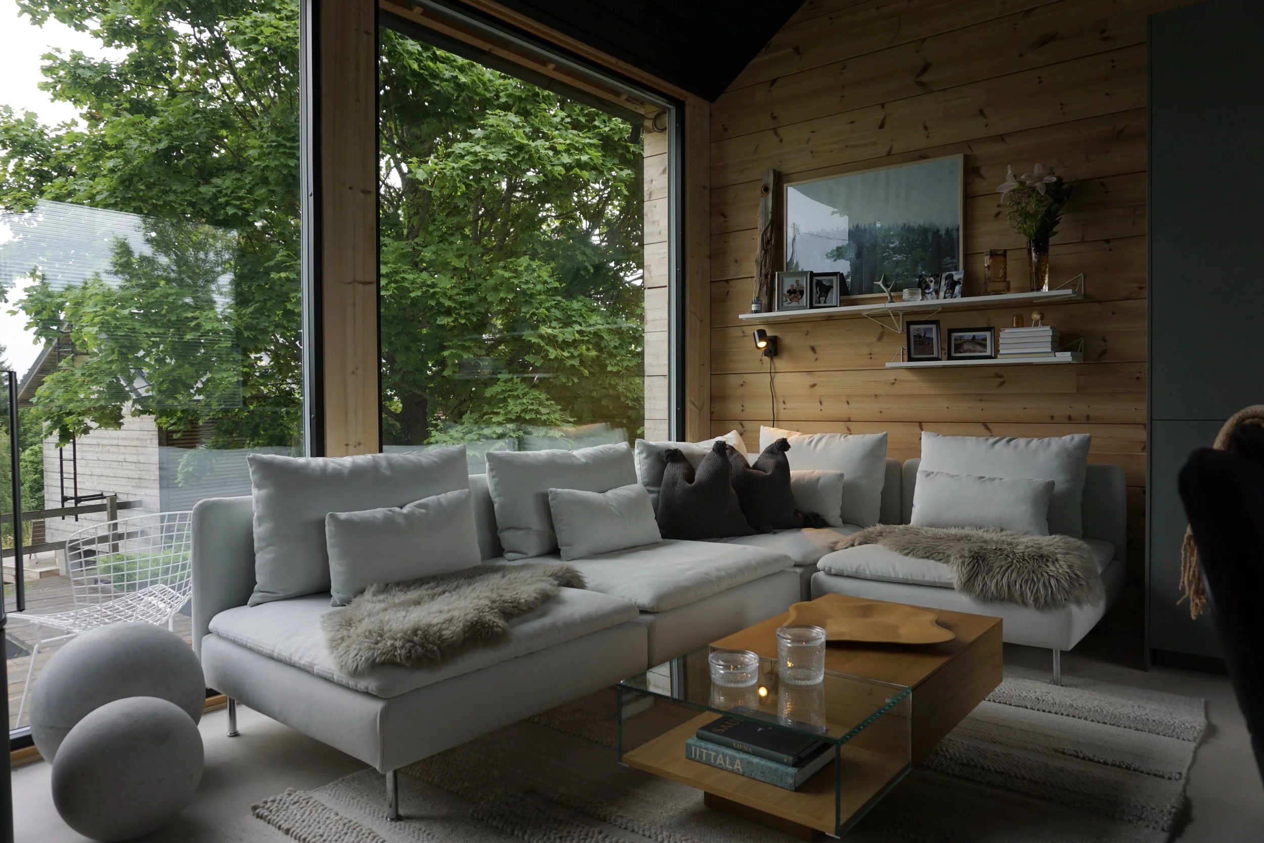 Living room with a large white sectional sofa with fur throws and cushions, glass coffee table with candles and books, wooden wall with framed photos and decor, large window revealing green trees outside.