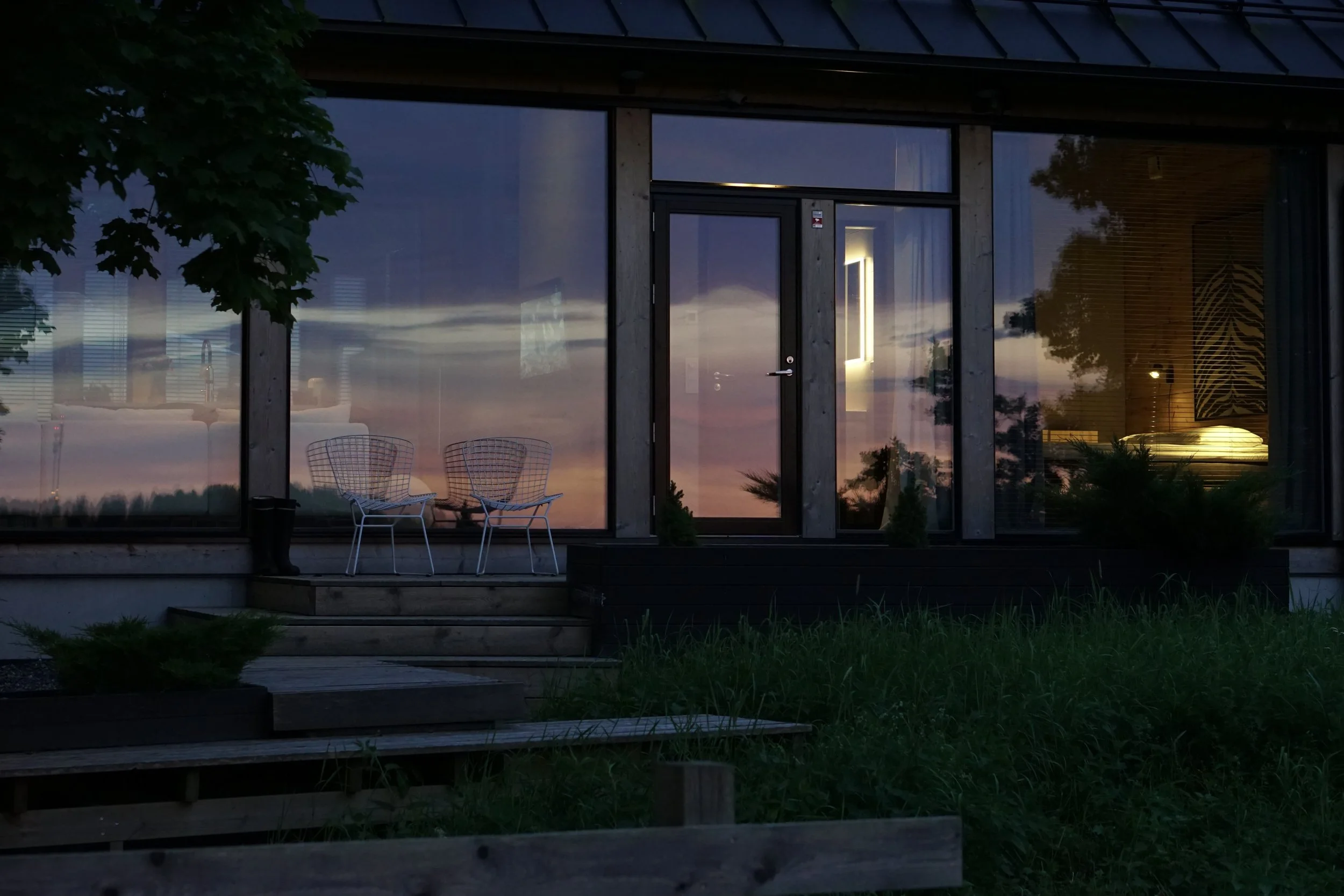A modern building with large glass windows reflecting the evening sky, featuring a lit interior visible through the glass, with two white wireframe chairs on a small wooden deck outside, surrounded by greenery and a tree on the left.