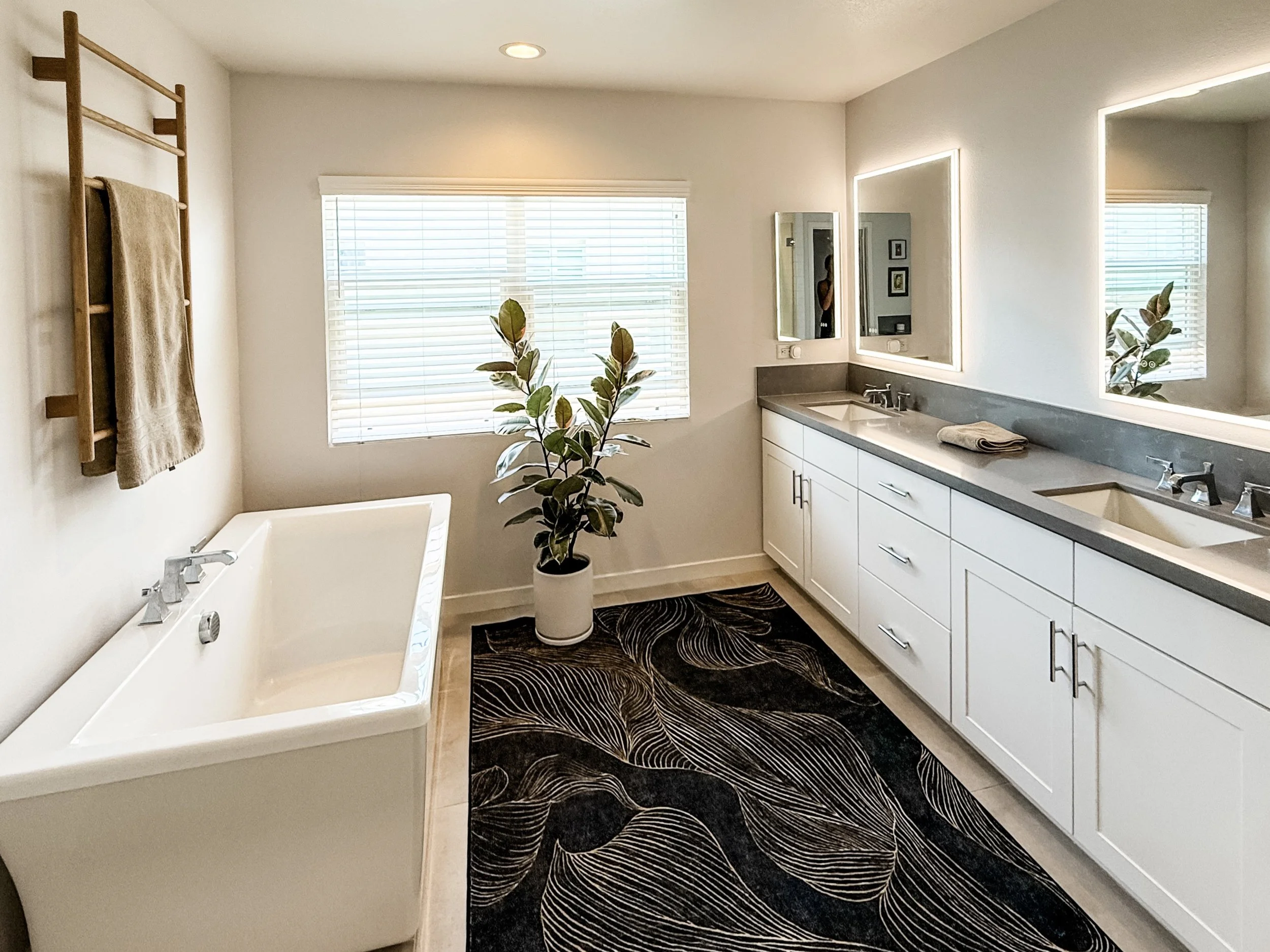 Bright bathroom with white vanity, black countertop, two illuminated mirrors, a window with blinds, a potted plant, a bath, and a towel rack on the wall.