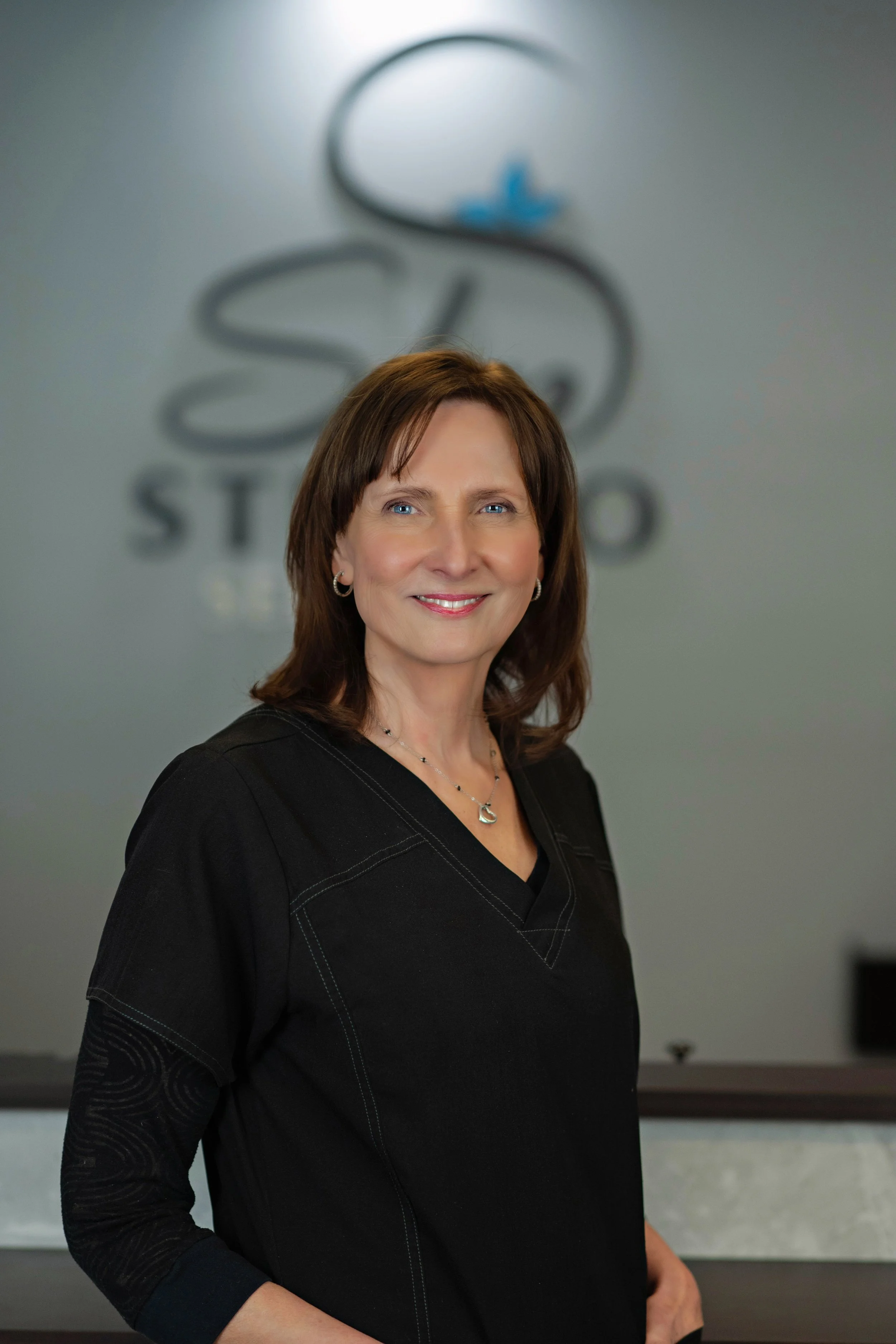 A smiling woman with shoulder-length brown hair wearing light blue scrubs, standing against a plain light blue background.