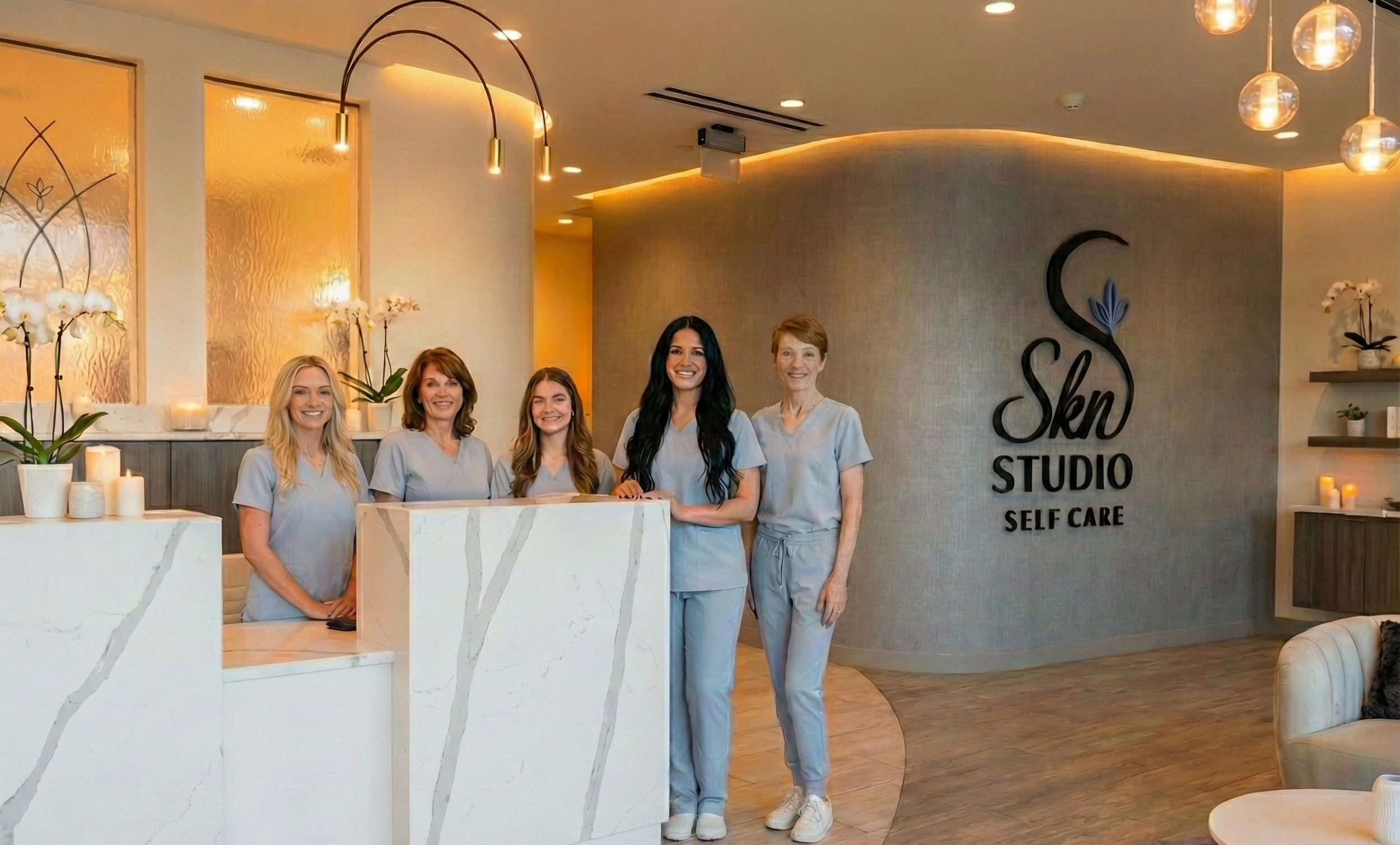 A group of five women in gray uniforms standing at the reception desk of a skincare studio, smiling. The background features a wall with a logo that says 'Skin Studio Self Care', decorated with orchids and candles.