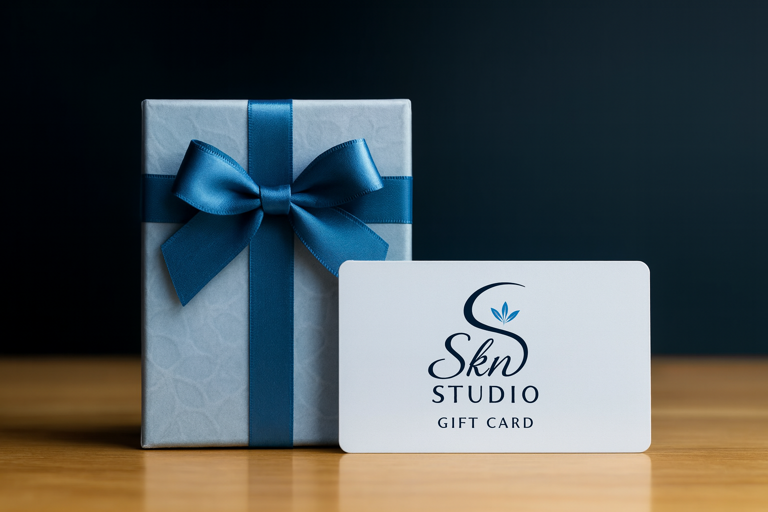 A wrapped gift box with a blue ribbon and bow next to a white gift card with the logo and text 'Skn Studio Gift Card' placed on a wooden surface against a dark background.