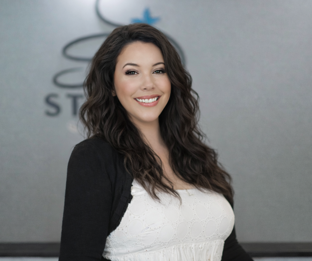 Professional woman with long dark hair smiling, wearing a light blue blazer over a matching top, standing against a neutral gray background.