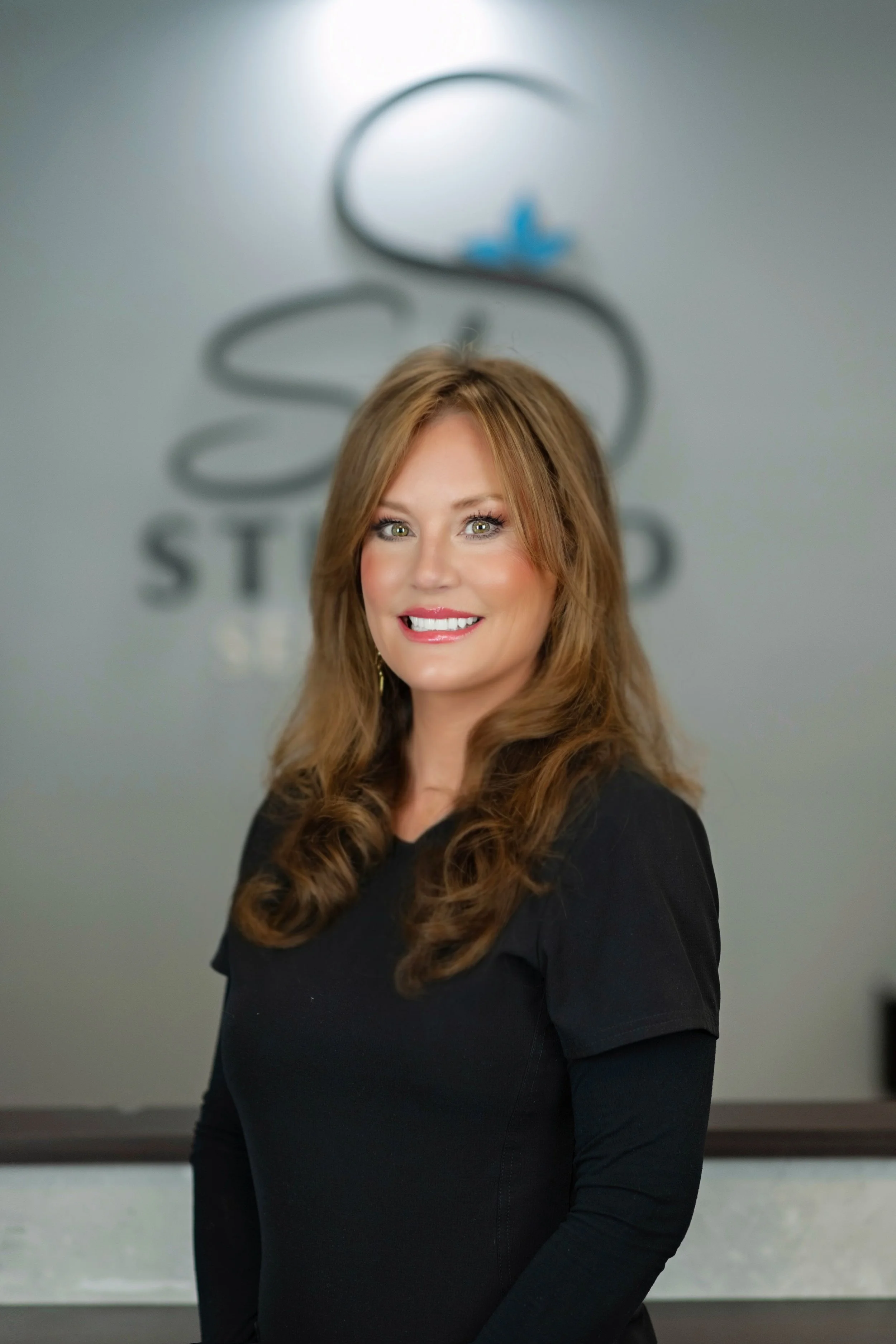 Portrait of a smiling woman with shoulder-length light brown hair, wearing blue medical scrubs against a plain gray background.