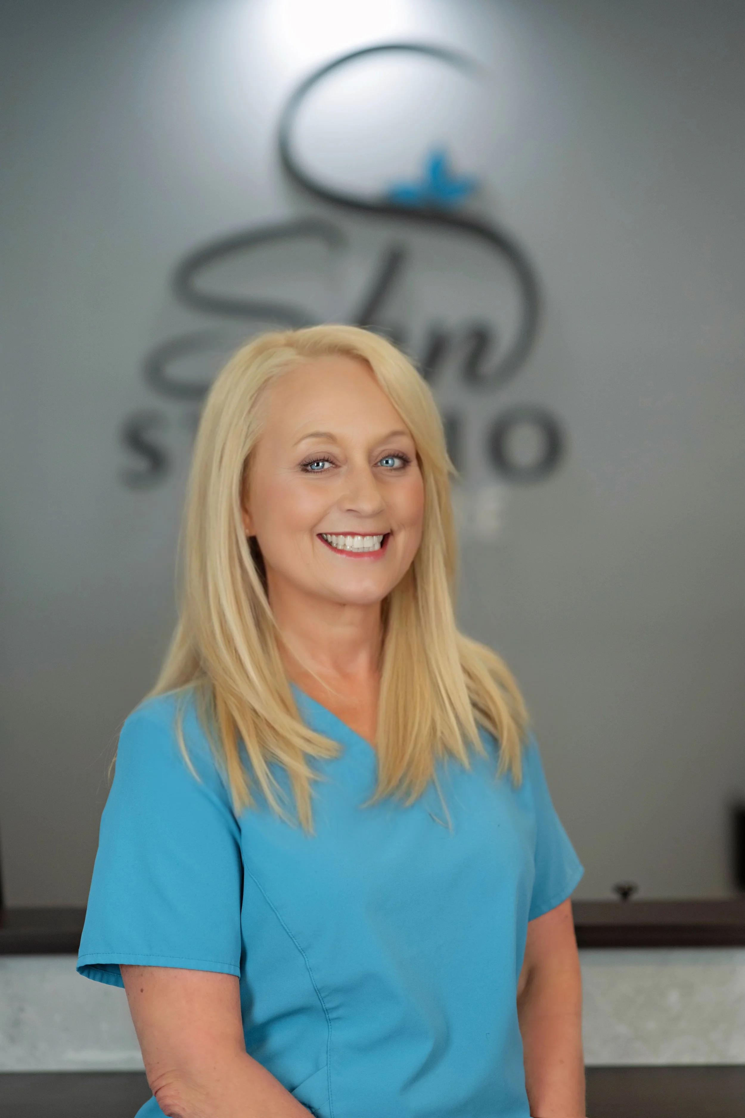 A woman wearing a blue medical scrub top, smiling, with blonde hair, earrings, and a neutral background.