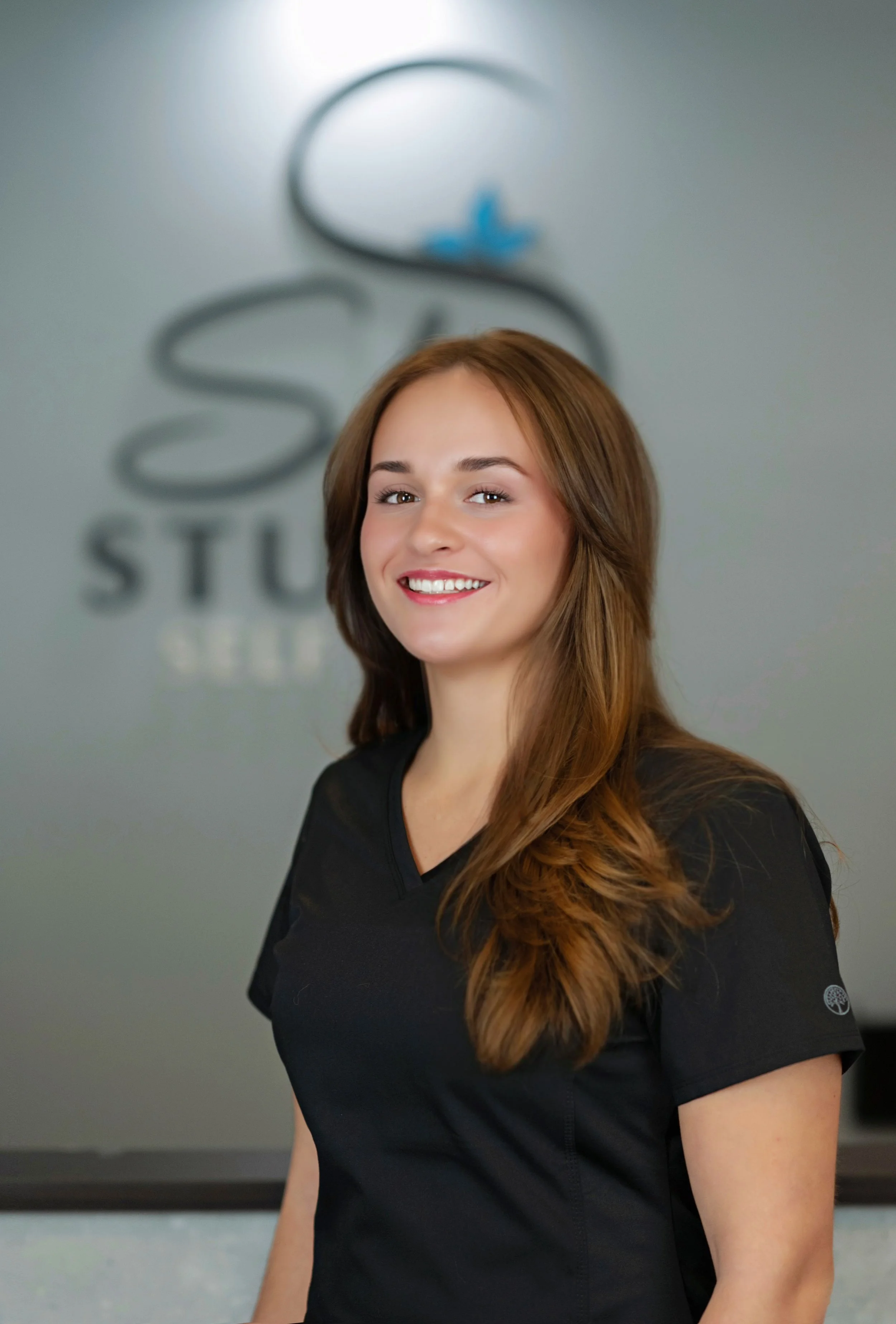 A young woman with shoulder-length brown hair wearing light blue medical scrubs, smiling in a clinical setting.