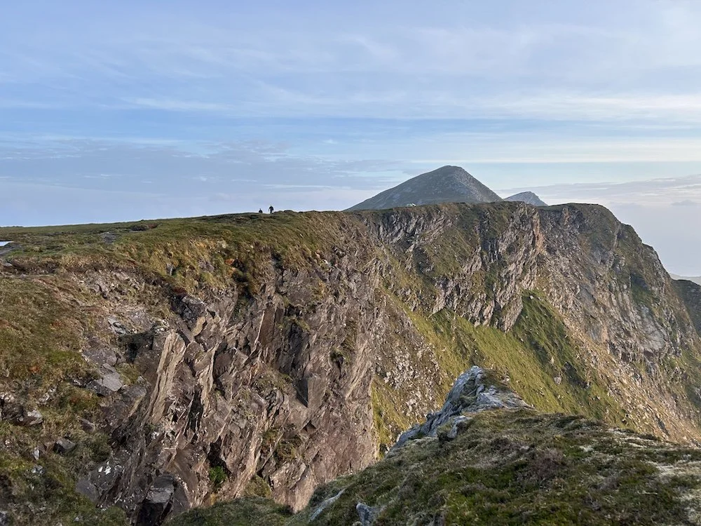Cliffside landscape with rugged rocks, patches of grass, a mountain in the background, and two small figures standing on the edge of the cliff under a partly cloudy sky.