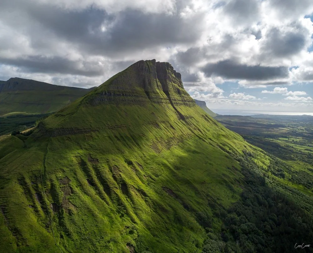 A lush green mountain with steep, layered slopes under a cloudy sky.