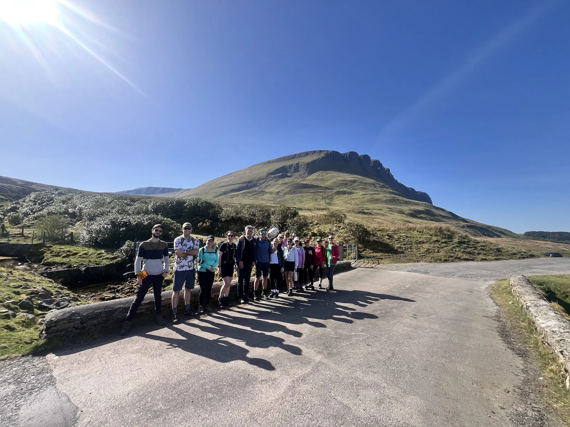 A group of people standing outdoors in front of a grassy hill with a mountain and clear blue sky in the background, with sunlight overhead.