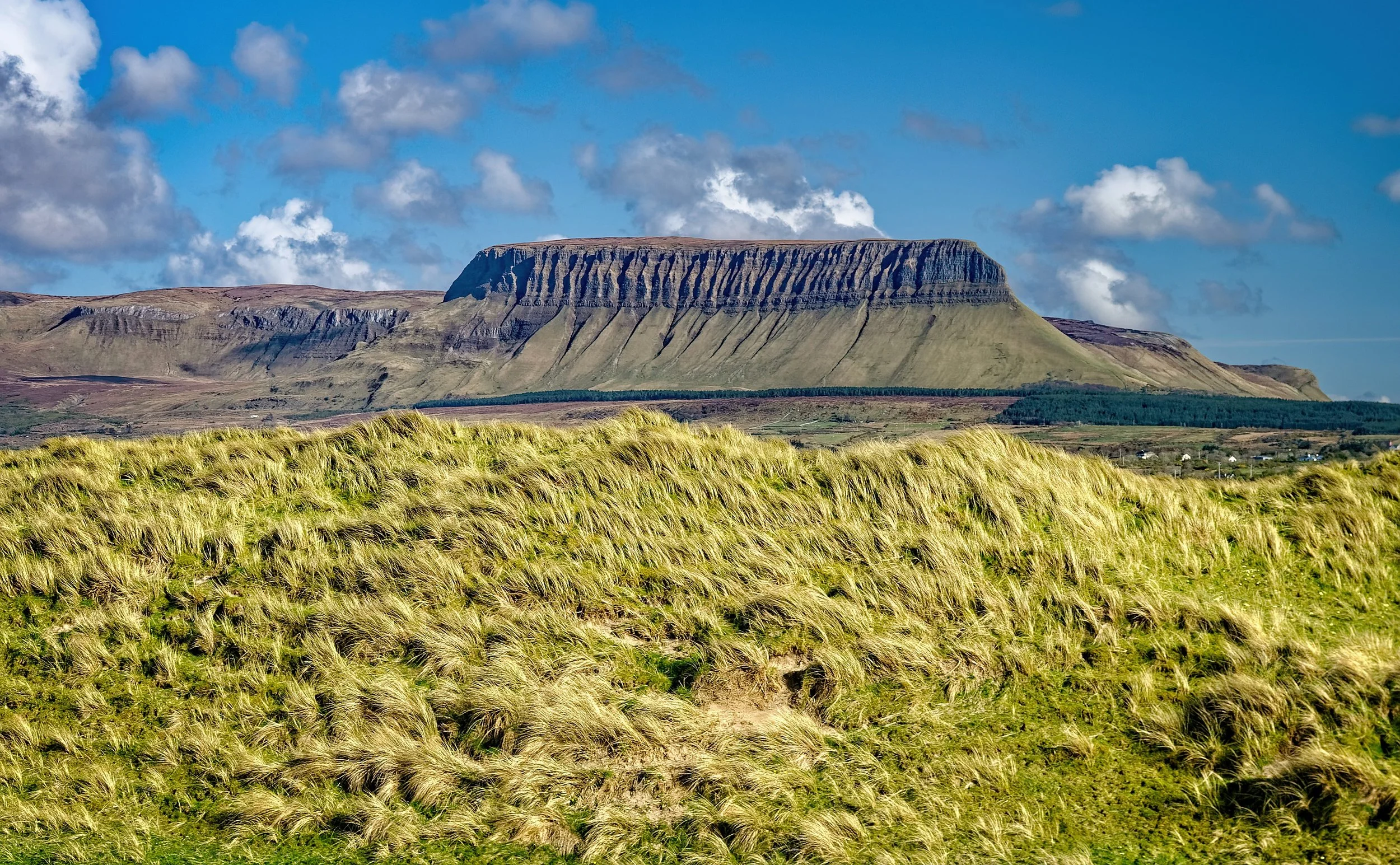 A large, flat-topped mountain with steep, layered slopes, surrounded by green grass and a partly cloudy sky.