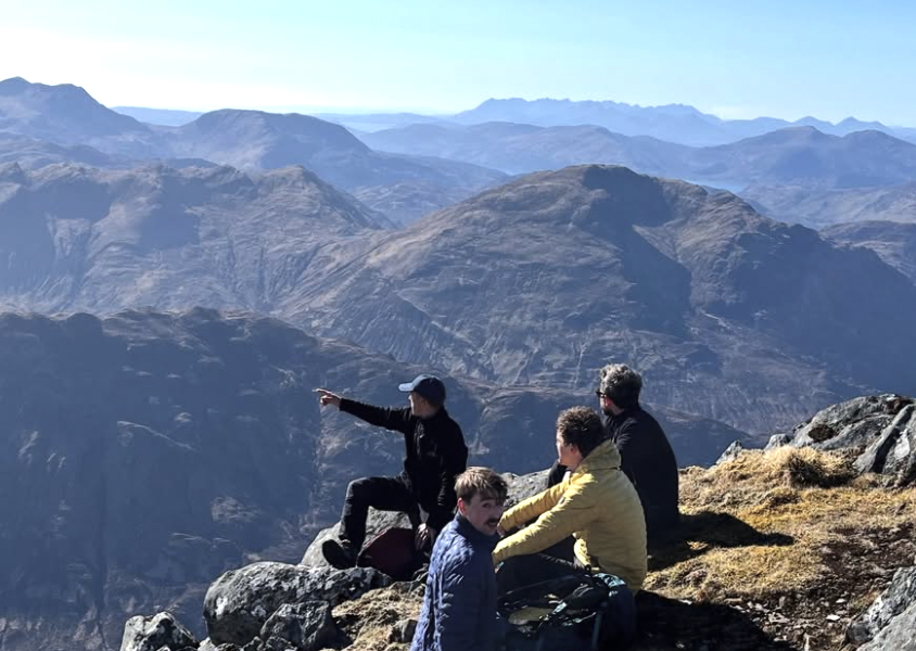 Four people sitting on rocks on a mountain, with a vast mountain range in the background, one person pointing at the scenery.