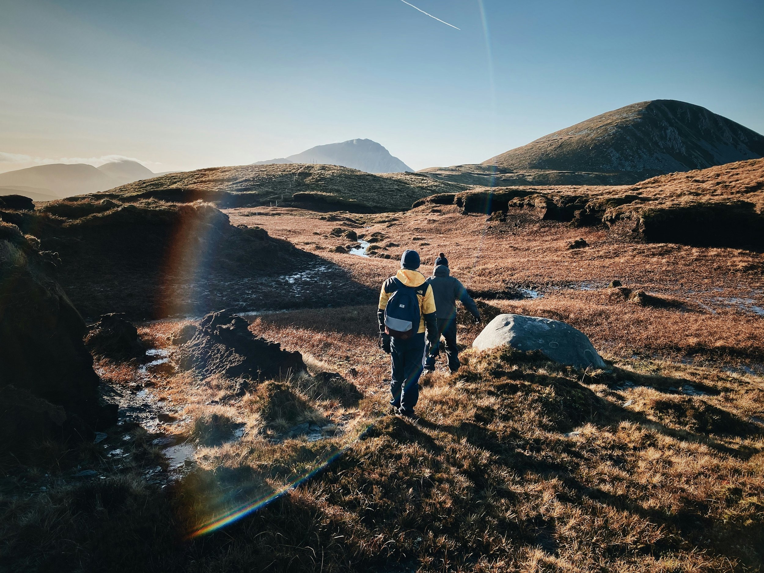Two hikers walking on a trail in a mountainous landscape with sunlight and clear sky.